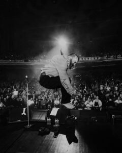 Black and white action shot of Teddy Swims leaping barefoot on stage during a live performance in front of a large, illuminated crowd.
