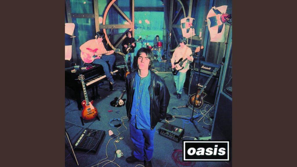 Overhead shot of the band Oasis in what appears to be a recording or rehearsal studio, with instruments like guitars, a piano, and drum kit visible. The band members are casually dressed, with Liam Gallagher looking directly at the camera. The 'Oasis' band logo is in the bottom right corner.